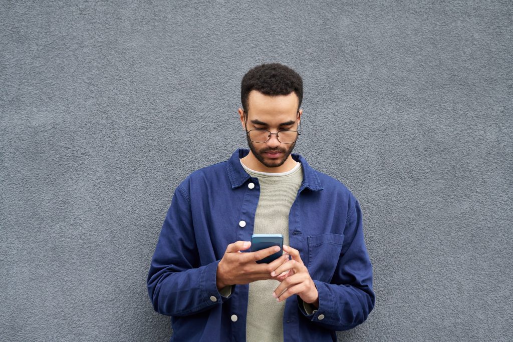 Young Black Man Browsing Smartphone While Standing Against Concrete Wall | Quest Mindshare Consumer taking an online survey, showing what drives survey respondent motivation
