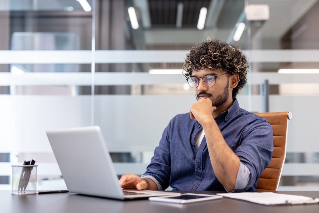 A thoughtful young Indian man is sitting at a desk in the office and looking at the laptop screen | Quest Mindshare Researcher reviewing survey data to learn how to prevent data fraudv