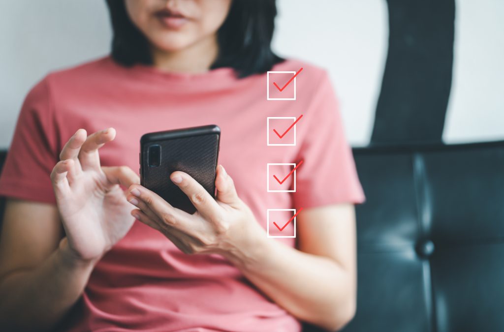 Woman holding smartphone and marking on checklist box with marker red Customer service feedback on mobile | Quest Mindshare Respondent taking a phone survey built with mobile-first survey design to protect survey data quality